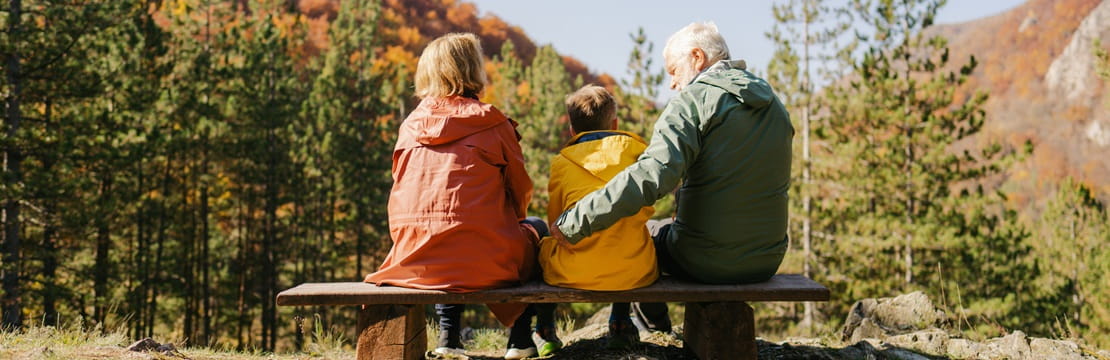 Grandparents sitting on a bench with their grandchild, overlooking a forested mountain with autumn colors under a clear sky.